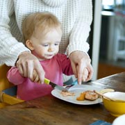 A mother helps their child learn to feed themselves.