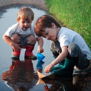 Young children floating home-made paper boats in a large puddle.